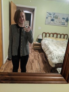 A young woman takes a selfie of herself in her bedroom mirror wearing a hand knit grey cardigan.