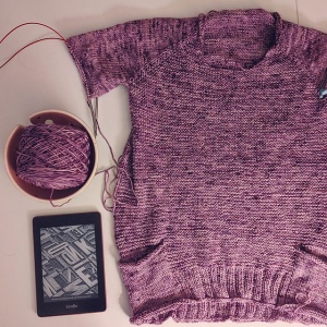 Top down view of a speckled knit sweater laying on a table with one of it's sleeves out of frame and the other in progress. Next to the sweater is a watermelon yarn bowl with a cake of yarn and an off kindle below.