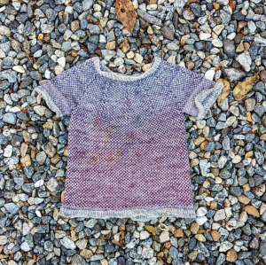 Close up of a sea glass mini toddler sweater, knit using a 1x1 colorwork pattern in a blue to purple gradient with a light gray yarn. The sweater has spots of green, red and yellow finger paint.